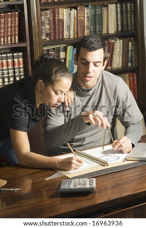 Young couple working at desk with a protractor, calculator, and ruler with books in the background. Vertically framed photo.