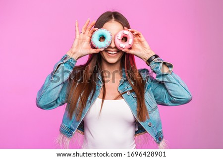 Studio positive photo young woman with sweets in jeans clothes indoors. Stylish funny girl model in blue denim fashionable jacket covers face with donuts near glamorous pink wall in the room.