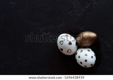 Three decorated and painted easter eggs lie in a black straw nest on a black background