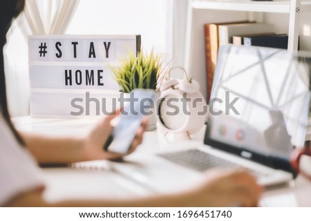 lightbox with text hashtag #STAYHOME glowing in lightand blurred woman working at home. Office worker on quarantine. Home working to avoid virus disease. Freelancer or remote worker concept. 