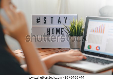 lightbox with text hashtag #STAYHOME glowing in lightand blurred woman working at home. Office worker on quarantine. Home working to avoid virus disease. Freelancer or remote worker concept. 
