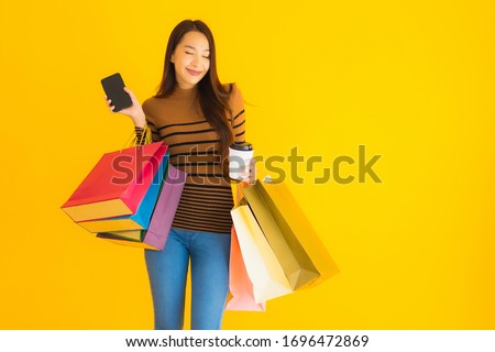 Beautiful young asian woman use smart mobile phone or cellphone with coffee cup and color shopping bag on yellow isolated background