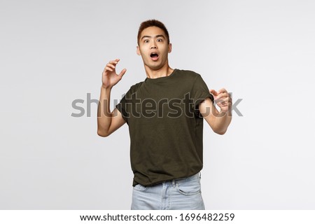 Portrait of alarmed and scared, shocked asian guy gasping, step back and raising hands up frightened, startled staring at something frightening, standing in awe over grey background Royalty-Free Stock Photo #1696482259