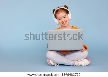 Full body positive little girl in headphones smiling and watching cartoon on modern laptop while sitting crossed legged on blue background
