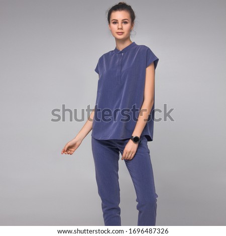 young stylish brunette nurse with long legs in blue medical costume is standing with one hand in air, looking straight and smiling cute on white wall background. medical concept. free space
