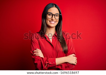 Young beautiful brunette woman wearing casual shirt and glasses over red background happy face smiling with crossed arms looking at the camera. Positive person.