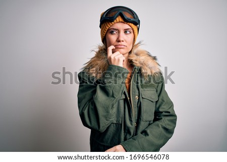 Young beautiful skier woman with blue eyes wearing snow sportswear and ski goggles with hand on chin thinking about question, pensive expression. Smiling with thoughtful face. Doubt concept.