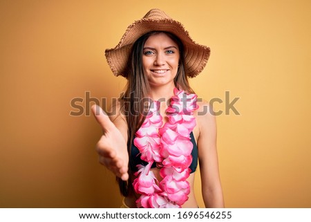 Young beautiful woman with blue eyes on vacation wearing bikini and hawaiian lei smiling friendly offering handshake as greeting and welcoming. Successful business.