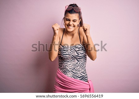 Young beautiful brunette woman on vacation wearing swimsuit over pink background excited for success with arms raised and eyes closed celebrating victory smiling. Winner concept.