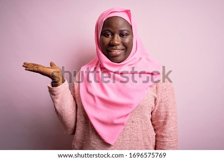 Young african american plus size woman wearing muslim hijab over isolated pink background smiling cheerful presenting and pointing with palm of hand looking at the camera.