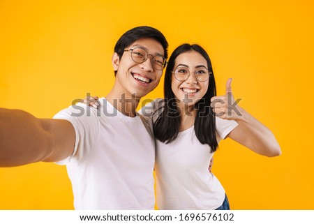 Image of joyful multinational couple taking selfie and gesturing peace sign isolated over yellow background