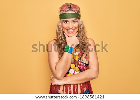 Beautiful blonde hippie woman wearing sunglasses and accessories over yellow background looking confident at the camera smiling with crossed arms and hand raised on chin. Thinking positive.