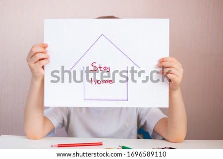 A house painted on a white sheet of paper with the words "stay home." The child holds a poster with an appeal. Children's creativity in quarantine.
