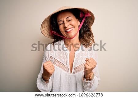 Middle age brunette woman wearing asian traditional conical hat over white background very happy and excited doing winner gesture with arms raised, smiling and screaming for success. Celebration 