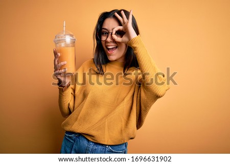 Young brunette woman drinking fresh orange juice from take away bottle over yellow background with happy face smiling doing ok sign with hand on eye looking through fingers