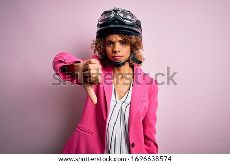 African american motorcyclist woman with curly hair wearing moto helmet over pink background looking unhappy and angry showing rejection and negative with thumbs down gesture. Bad expression.