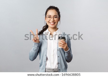 Office lifestyle, business and people concept. Optimistic, friendly-looking asian girl show peace sign, smiling kawaii, enjoying morning fresh cup coffee from local cafe, stand grey background