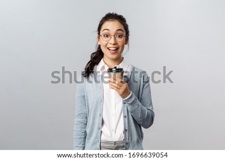 Lifestyle, cafe and people concept. Amused, excited smiling asian woman looking with interest and amazement at camera, open mouth fascinated, holding cup of take-away coffee, stand grey background