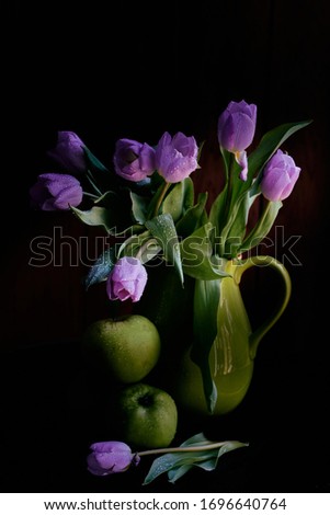 A bouquet of lilac tulips in a green jar with green apples covered with raindrops on black background