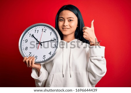 Young beautiful chinese woman holding big clock standing over isolated red background happy with big smile doing ok sign, thumb up with fingers, excellent sign