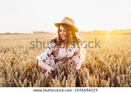 Young girl with long curly hairand freckles face, in hat, in light white dress with floral print, standing in wheat field, posing for camera, in background sundown