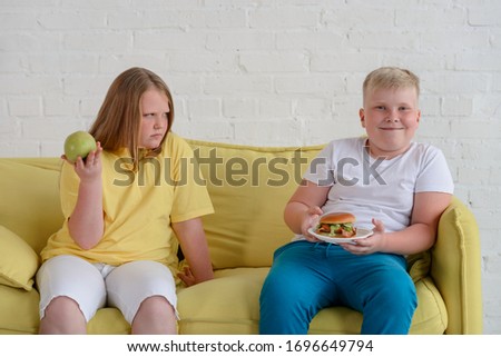 Fat kids eating different food. Girl is jealous of boy eating tasty hamburger while she's having an apple.