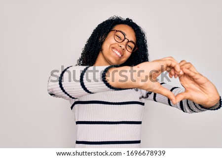 Young african american woman wearing striped sweater and glasses over white background smiling in love doing heart symbol shape with hands. Romantic concept.