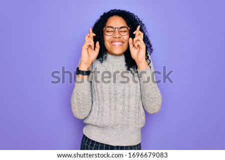 Young african american woman wearing casual sweater and glasses over purple background gesturing finger crossed smiling with hope and eyes closed. Luck and superstitious concept.
