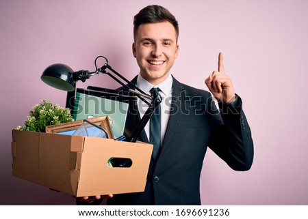 Young handsome caucasian business man holding cardboard box unemployment fired from job surprised with an idea or question pointing finger with happy face, number one