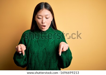 Young beautiful asian woman wearing green winter sweater over yellow isolated background Pointing down with fingers showing advertisement, surprised face and open mouth