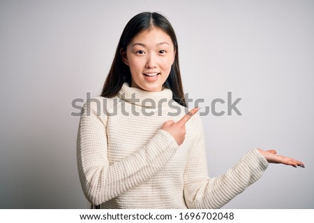 Young beautiful asian woman wearing casual sweater standing over isolated background amazed and smiling to the camera while presenting with hand and pointing with finger.