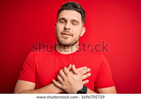 Young man with blue eyes wearing casual t-shirt over red isolated background smiling with hands on chest with closed eyes and grateful gesture on face. Health concept.