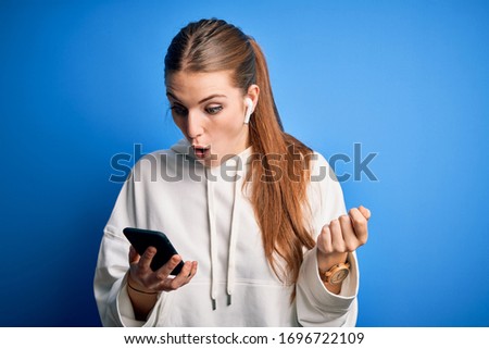 Young redhead sportswoman doing sport listening to music using earphones and smartphone Pointing down with fingers showing advertisement, surprised face and open mouth
