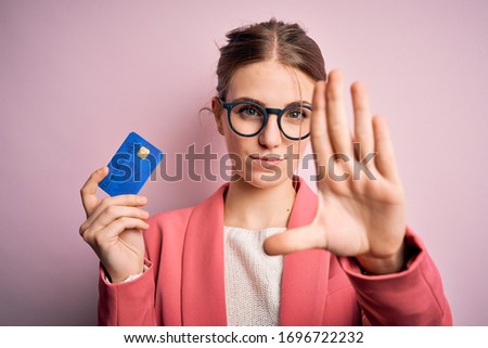 Young beautiful redhead woman holding credit card over isolated pink background with open hand doing stop sign with serious and confident expression, defense gesture