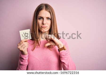 Young beautiful redhead woman holding reminder paper with smile emoji message with angry face, negative sign showing dislike with thumbs down, rejection concept