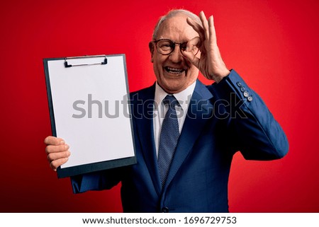 Senior grey haired business man holding clipboard over red background with happy face smiling doing ok sign with hand on eye looking through fingers