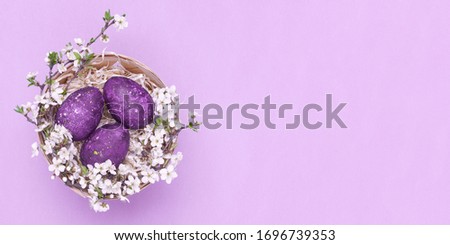 Violet easter eggs in a basket with flowers on a violet background. Horizontal photograph. Top view Flatley. Top view. Object on the left side of the photo