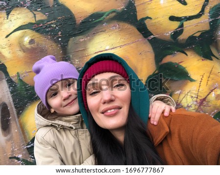 a young beautiful mother and little daughter are smiling at the camera nearby, in the background on the wall there is a bright picture