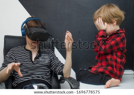 A young father plays in a virtual reality helmet. Little boy son is sitting nearby and crying. Child needs attention from his father.