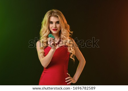 Blonde girl in red fitting dress and necklace. She smiling, showing two green chips, posing on colorful background. Poker, casino. Close-up