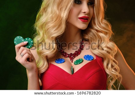 Blonde girl in red dress. Smiling, showing two green chips and some colorful are on her breasts. Colorful smoky background. Poker, casino. Close-up