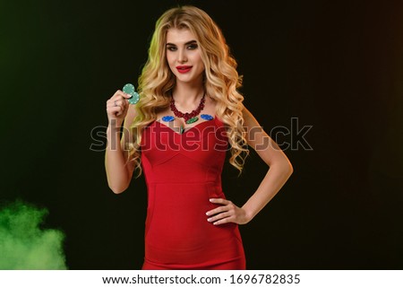 Blonde female in red dress. Smiling, showing two green chips and some colorful are on her breasts. Colorful smoky background. Poker, casino. Close-up