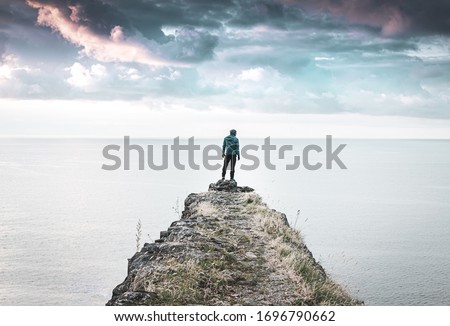 Person stands alone and looks around on the edge of  the cliff with vast sea and dramatic sky in the background.  Concept of solitude, adventuree and lifestyle. 2020 Royalty-Free Stock Photo #1696790662