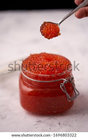 Hand with a spoon with red caviar over a glass jar filled with red caviar of salmon fish on a light background. Seafood, caviar, healthy food, top view                              