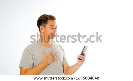 Calm young asian male in grey t-shirt isolated on white background looking his phone and smiling, male hold phone on chest close to heart feel grateful after seeing notification on her mobile phone