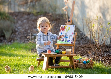 Sweet toddler child, painting eggs in garden with little chicks running around him on sunset