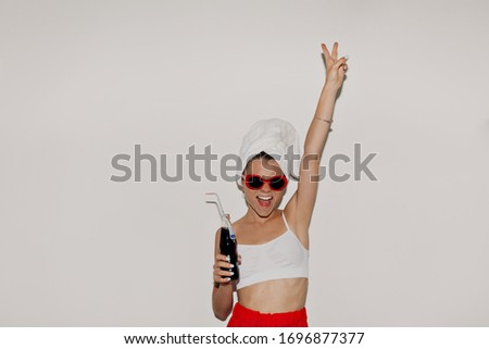 Blissful european girl in white top and towel on the head and red sunglasses holding up hand and smiling on isolated background. Adorable girl posing with happy face expression.