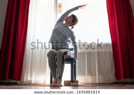 Young cheerful woman practicing yoga, sitting on chair doing side bend exercise, working out at the living room at home Royalty-Free Stock Photo #1696881814