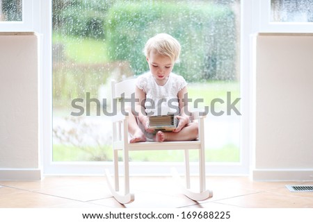 Adorable little blonde toddler girl sitting cozy in a white rocking chair next to a big window on a rainy day looking pictures in old family photo album