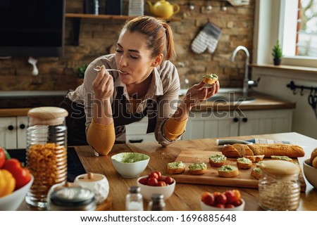 Smiling woman enjoying in taste of healthy food while making avocado bruschetta in the kitchen.  Royalty-Free Stock Photo #1696885648